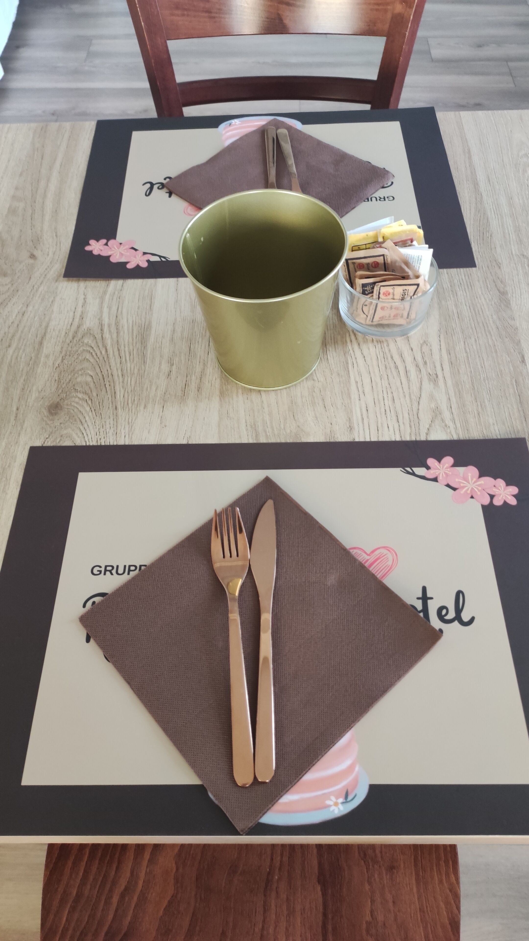 A dining table with a placemat, a green mug, a glass container with snacks, and cutlery.