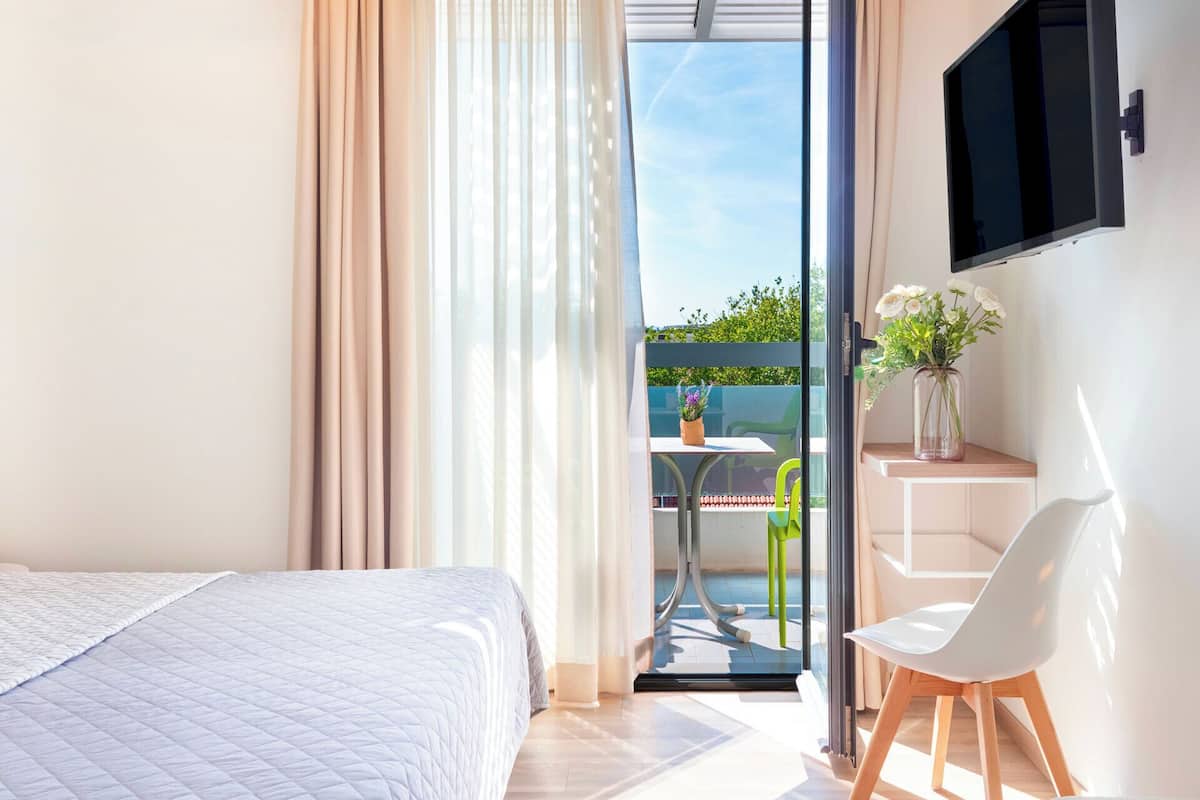 Un dormitorio con cama, un escritorio con silla, televisión y balcón con mesa y sillas.