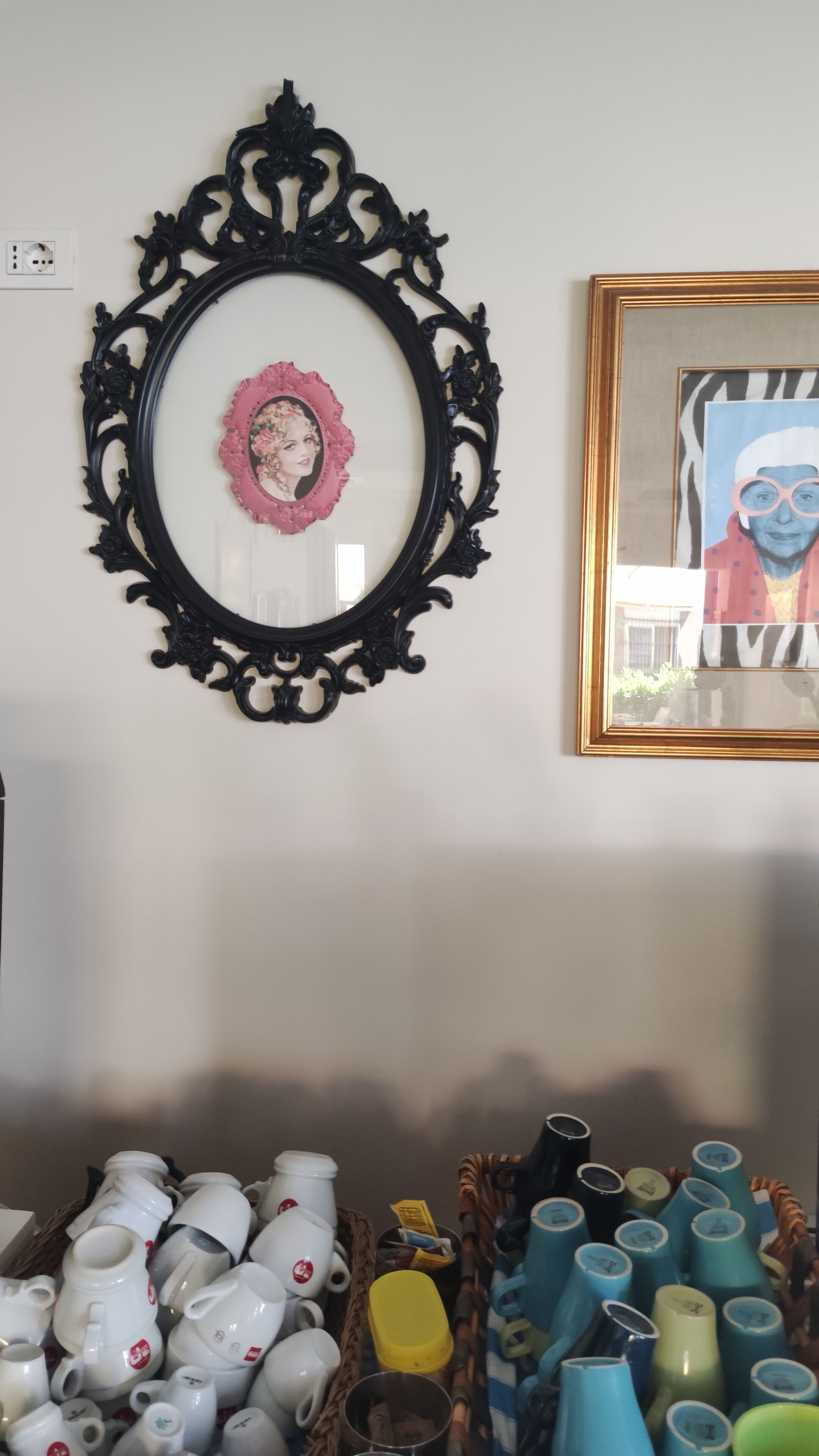 Un mur orné d’un cadre noir décoratif, sur lequel sont accrochés une photo encadrée en rose, une œuvre d’art encadrée et un panier rempli de tasses en céramique.