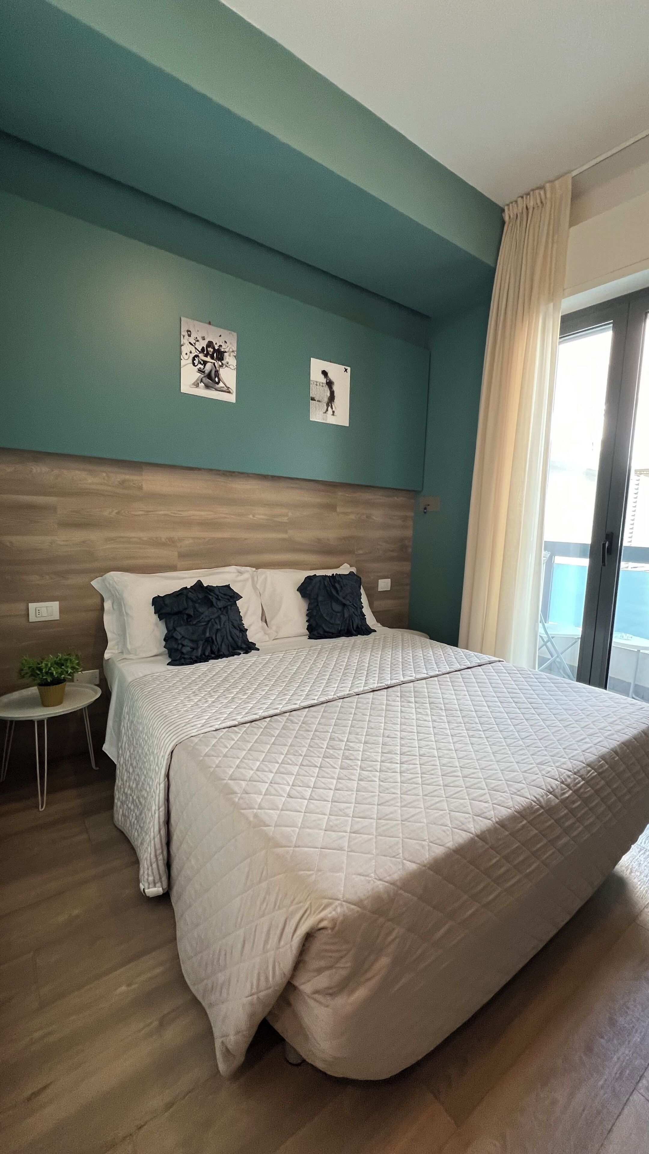 A bedroom with a large bed, a wooden headboard, teal walls, and a small side table with a plant.