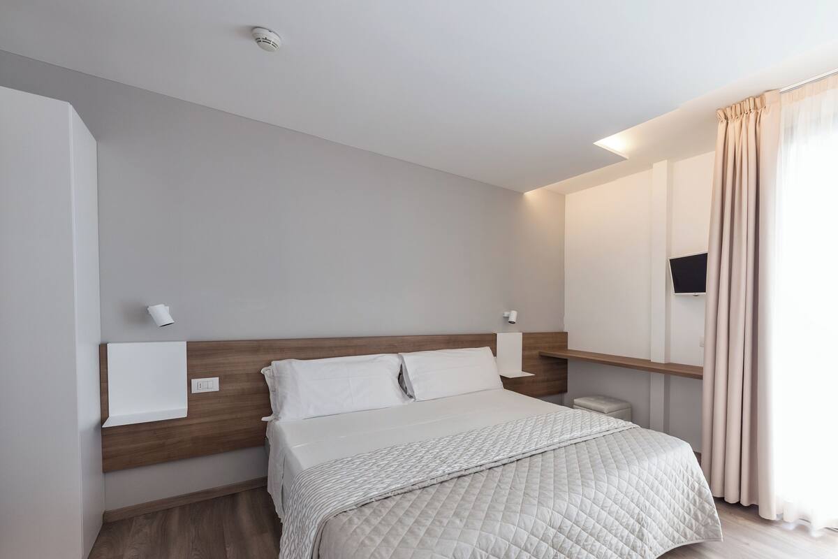 Una habitación de hotel moderna con una cama grande, cabecero de madera, mesitas de noche, un televisor de pantalla plana y una ventana con cortinas.