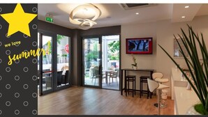 Ein modernes Interieur mit Glastür, Deckenleuchte, Wand-TV und einer Rezeption mit Sitzgelegenheiten.