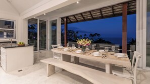 A modern dining area with a large table set for six, a kitchen area, and a view of the ocean at dusk.