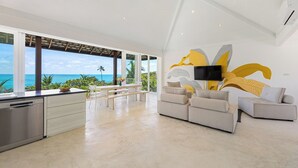 Un amplio salón con un gran ventanal con vistas al océano, una cocina moderna con lavavajillas y un televisor de pantalla plana grande montado en la pared.