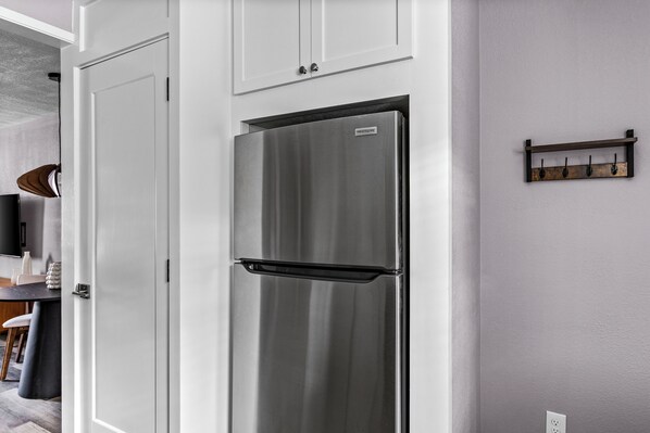 A stainless steel refrigerator in a kitchen with white cabinets and a wooden shelf with hooks.
