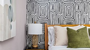 Una cama bien hecha con un cabecero de madera, una mesita de noche con una lámpara, y una pared con un papel tapiz estampado con motivos geométricos.