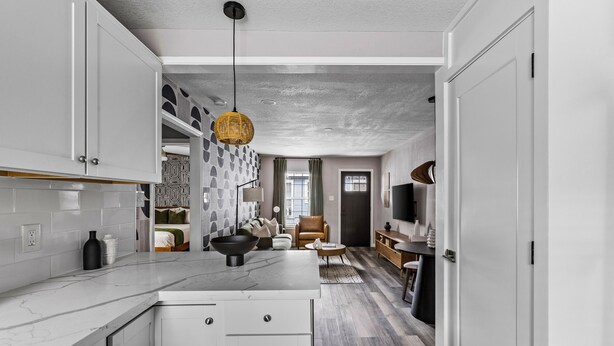 Una cocina moderna con armarios blancos, encimera de mármol y una isla central. Hay un rincón de comedor con un sofá y un televisor.