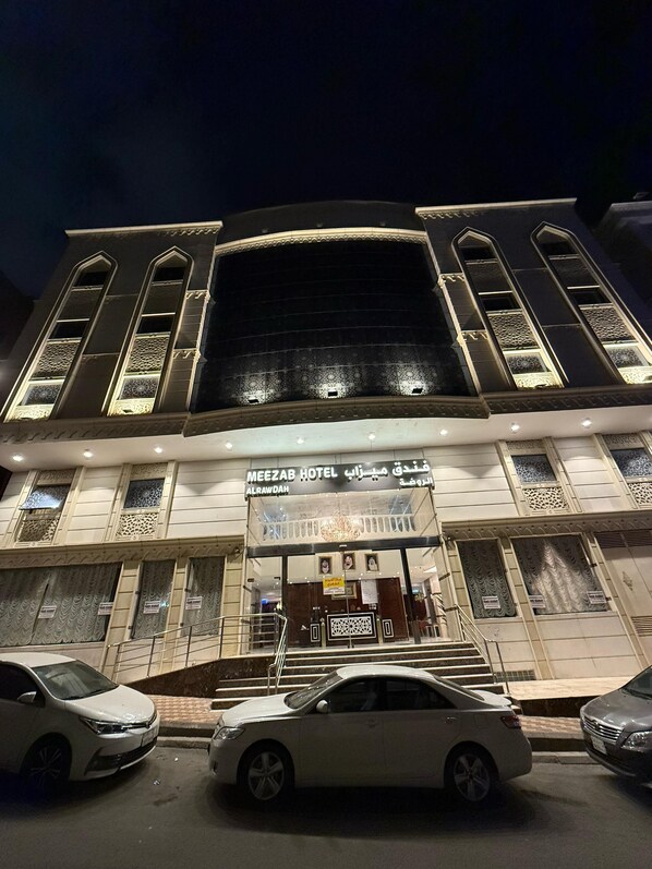 A hotel with a large arched entrance, multiple balconies with intricate railings, and a sign indicating it is the Meezab Hotel in Al Rawdah.