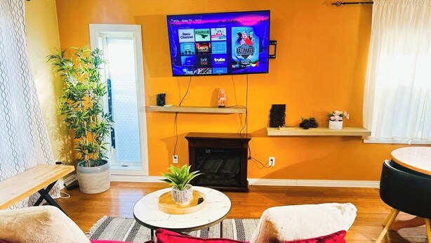 A living room with a TV mounted on the wall, a fireplace, a sofa, and a small table with a plant. - Cozy 2BER 1.5 Bath Home Near Rogers/2min walk 2 Stadium/RAH /NAIT/FastWIFI/Parkx (Edmonton)