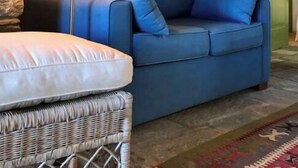 Ein Wohnzimmer mit blauem Sofa, einem Korbstuhl, einer Stehlampe und einem Gemälde an der Wand.