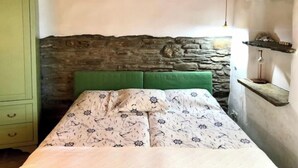 Ein Schlafzimmer mit einer Steinwand, einem mit grünem Stoff bezogenen Kopfteil, einer gemusterten Tagesdecke und einem hölzernen Regal mit einer Lampe.