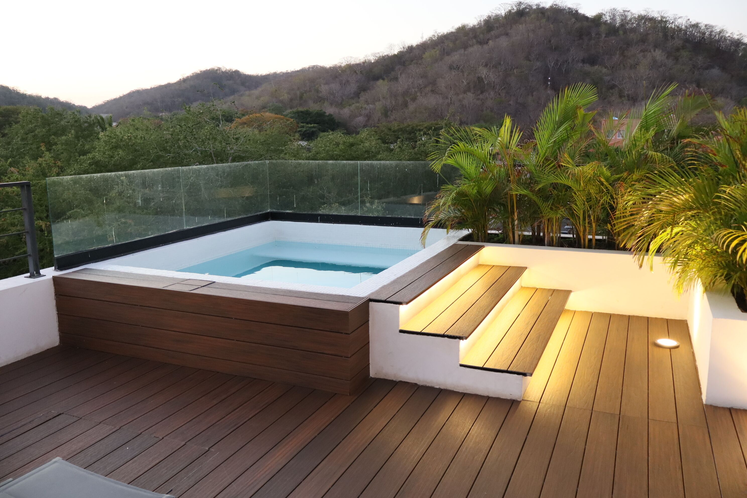 Un bain à remous sur une terrasse en bois dotée d’une rambarde en verre, entouré de verdure et de collines.