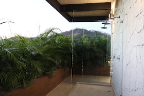 A modern shower enclosure with a glass door, surrounded by lush greenery and a view of a hilly landscape.