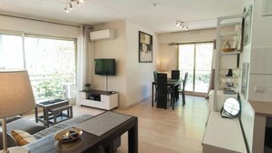 A modern living room with a sofa, coffee table, and TV. There is a dining area with a table and chairs. The room has large windows and a ceiling fan. - Elegant Apt w/2 Terraces/Mins to Beach & Shops (Antibes)