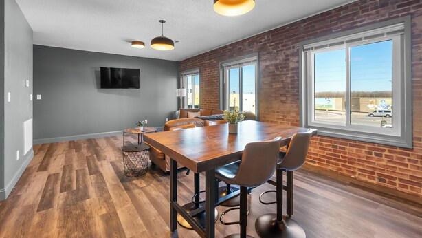 Une pièce spacieuse avec une grande table en bois, plusieurs chaises et un mur en briques. Deux grandes fenêtres offrent une vue sur l’extérieur.