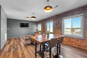A spacious room with a large wooden table, multiple chairs, and a brick wall. There are two large windows providing a view of the outside.