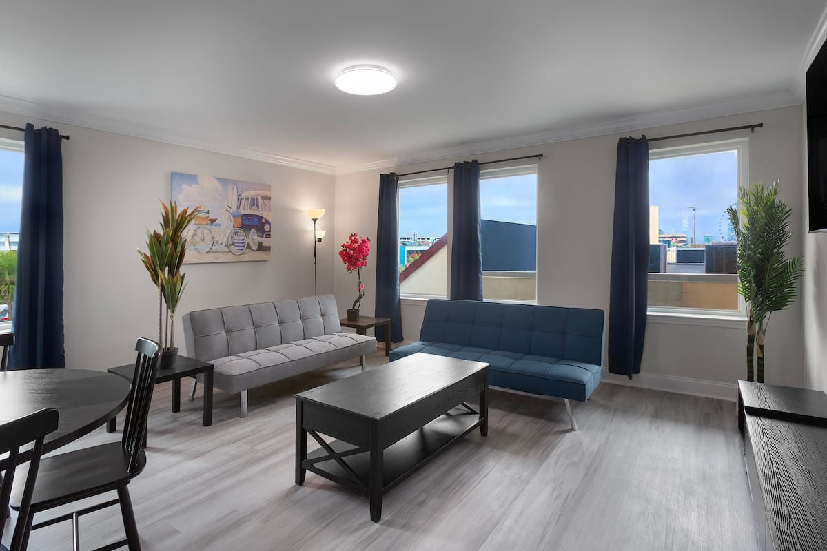 A modern living room with a grey sofa, blue sofa, and a black coffee table. There is a dining table with chairs, a potted plant, and a painting on the wall.