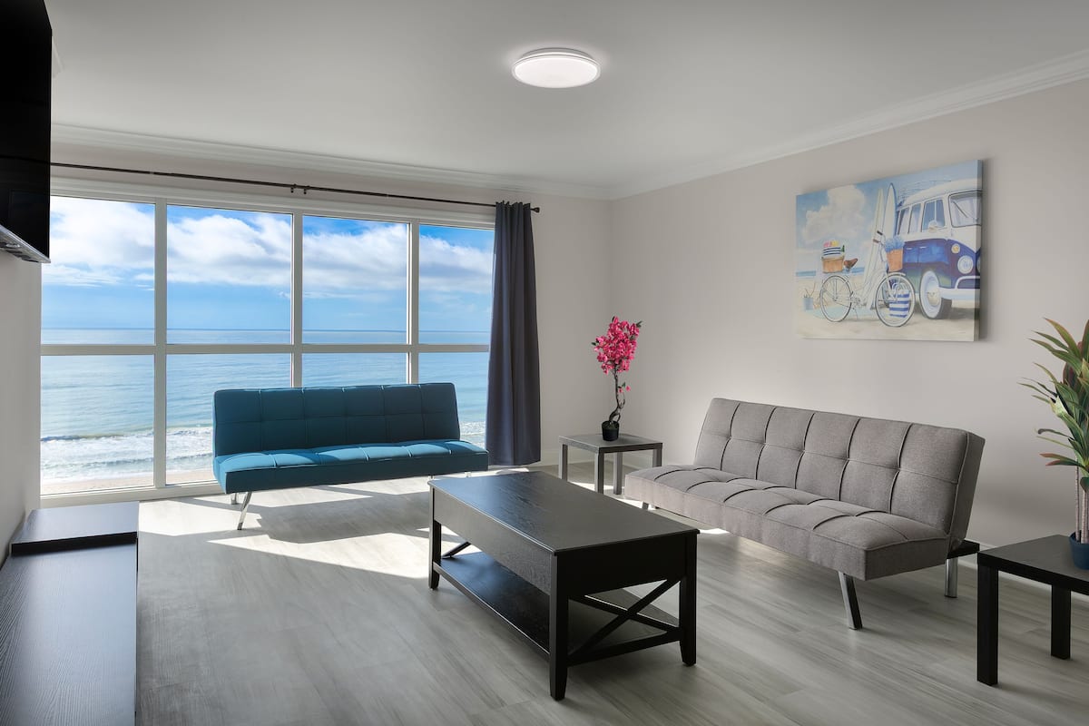 A modern living room with a large window overlooking the ocean, a blue sofa, a grey sofa, a black coffee table, and a painting of a van and surfboard on the wall.