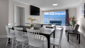 A dining area with a large table and white chairs, a flat-screen TV on the wall, and a view of the ocean through a large window. - Ocean Escape Boardwalk Oceanfront  (Myrtle Beach)