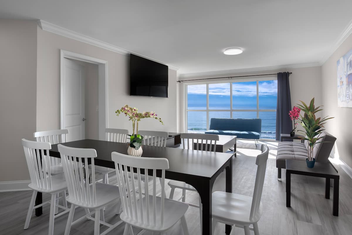 A dining area with a large table and white chairs, a flat-screen TV mounted on the wall, and a view of the ocean through a large window.