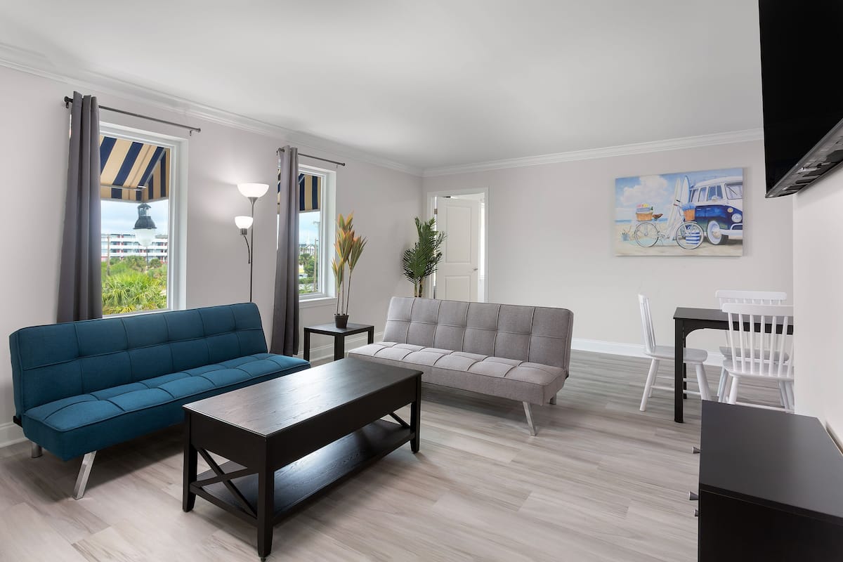 A modern living room with a blue sofa, a grey sofa, a wooden coffee table, and a dining area with white chairs and a small table.