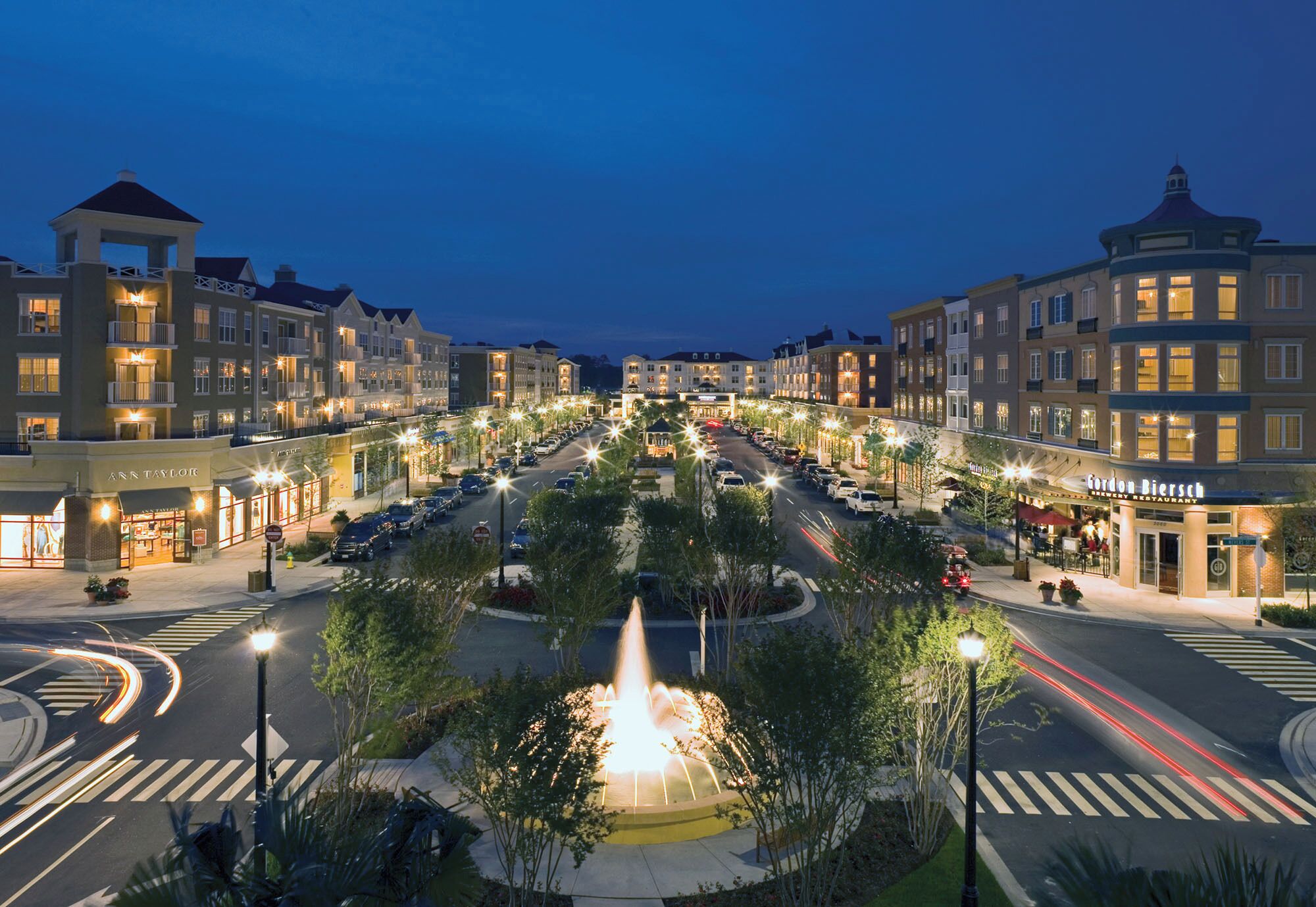 Una intersección urbana bien iluminada con una fuente central, rodeada de edificios de varios pisos que albergan comercios.