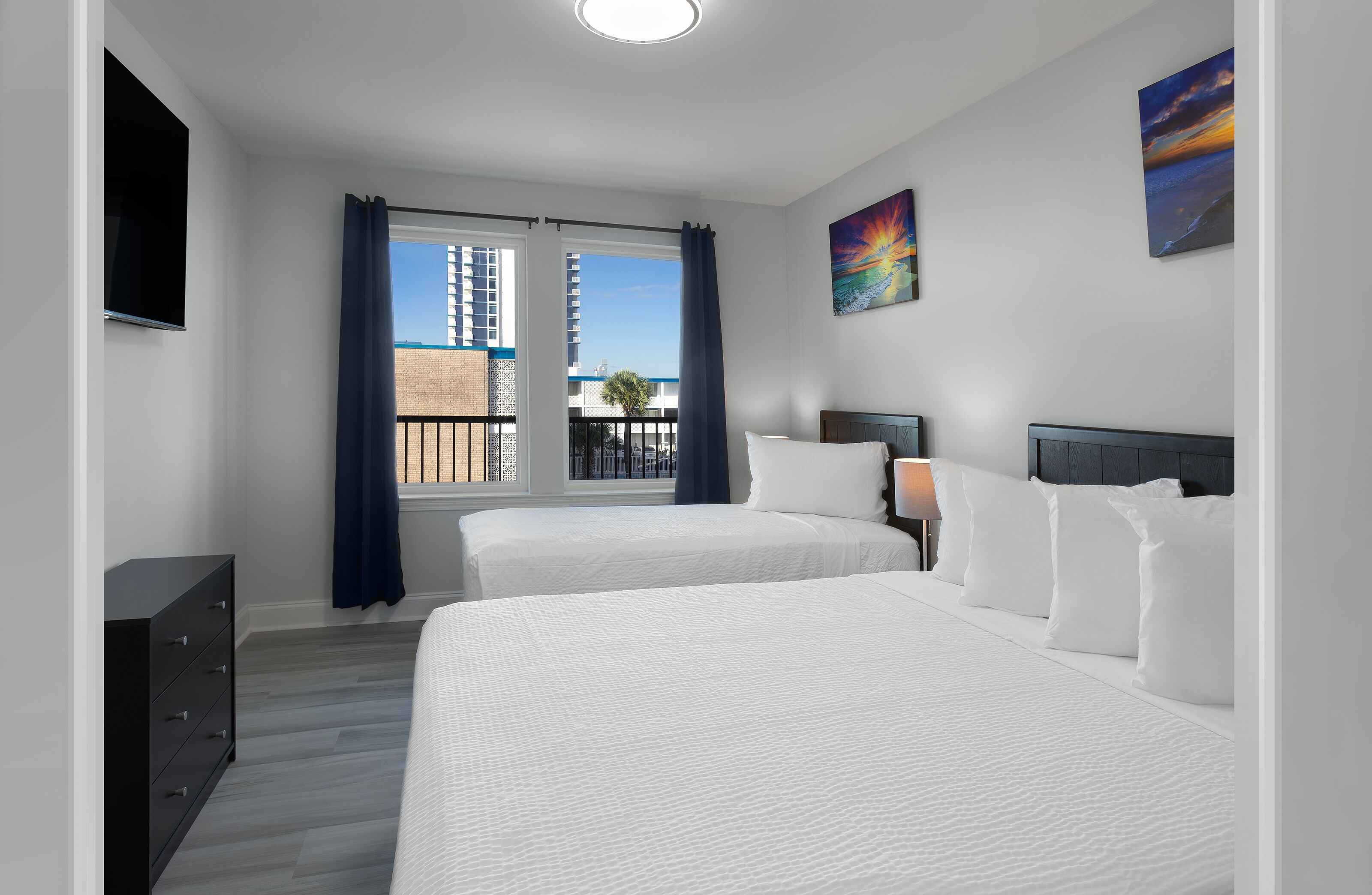 Ein Hotelzimmer mit zwei Betten, einem Fernseher, einem Fenster mit Vorhängen und Bildern an den Wänden.