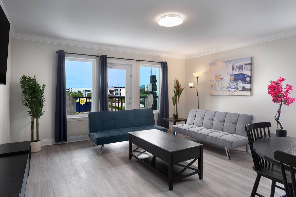 A modern living room with a blue sofa, grey sofa, and a dining table with chairs. There is a large window with curtains, a potted plant, and a painting on the wall.