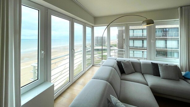 Een moderne woonkamer met een grote grijze bank, een vloerlamp en uitzicht op het strand en gebouwen door grote ramen.