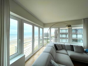 A modern living room with a large grey sofa, a floor lamp, and a view of the beach and buildings through large windows. - Apartment by Epernay Square With Sea View (Middelkerke)