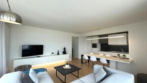 Een moderne woonkamer met een bank, salontafel en televisie. Er is een eethoek met tafel en stoelen. De kamer heeft een minimalistisch design met witte muren en een grote spiegel.
