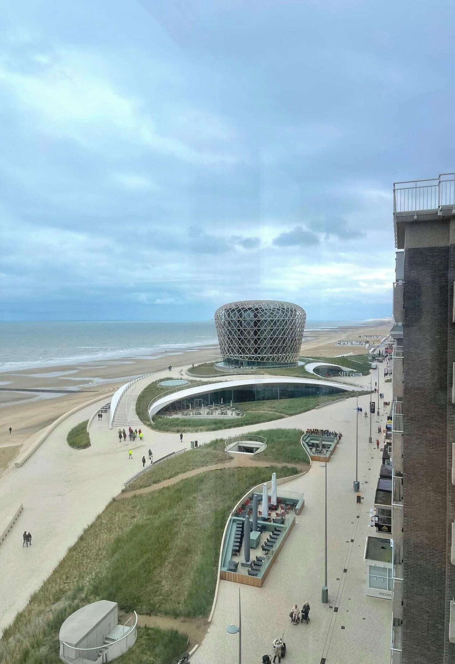 A modern building with a unique spherical design, situated near a beach with a promenade and a parking area.