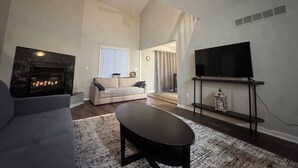 Un salon avec une cheminée, un canapé, une table basse, un téléviseur posé sur un meuble et une vue sur une autre pièce.