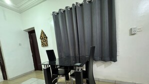 Un coin repas avec une table en verre et des chaises noires, un rideau gris et un sol carrelé.