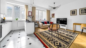 Um apartamento moderno com cozinha, sala de jantar e sala de estar. Possui uma janela grande com persianas, uma televisão e uma luminária decorativa no teto.