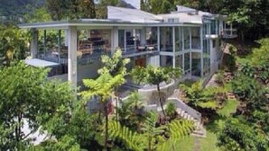 Une maison moderne en verre, dotée d’un intérieur spacieux, entourée d’une végétation luxuriante et offrant une vue sur un paysage montagneux.
