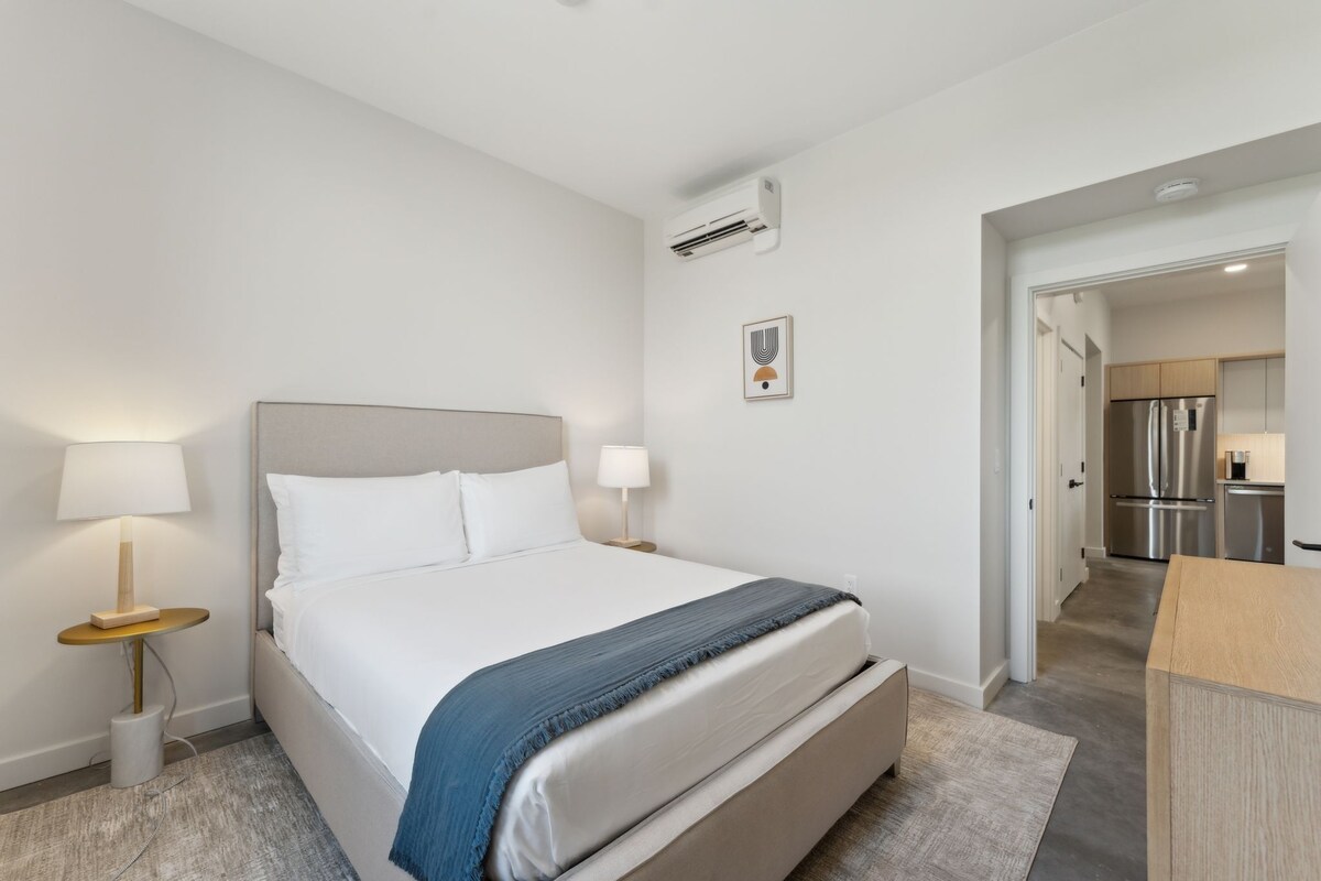 Ein modernes Hotelzimmer mit einem großen Bett, Nachttischlampen, einem kleinen Tisch und Blick in den Küchenbereich.
