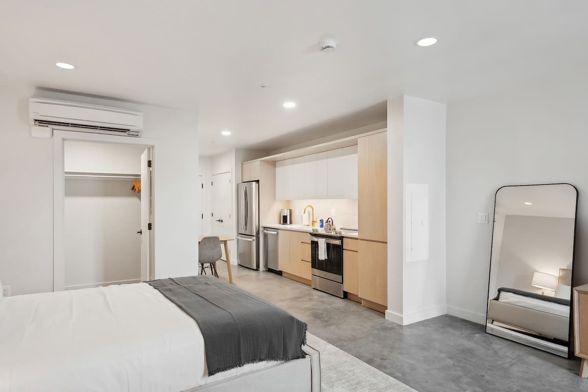 A modern hotel room with a bed, a desk, a kitchenette, and a large mirror.