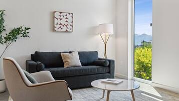 A modern living room with a grey sofa, a beige armchair, a round coffee table, a floor lamp, and a large window offering a view of greenery and mountains.