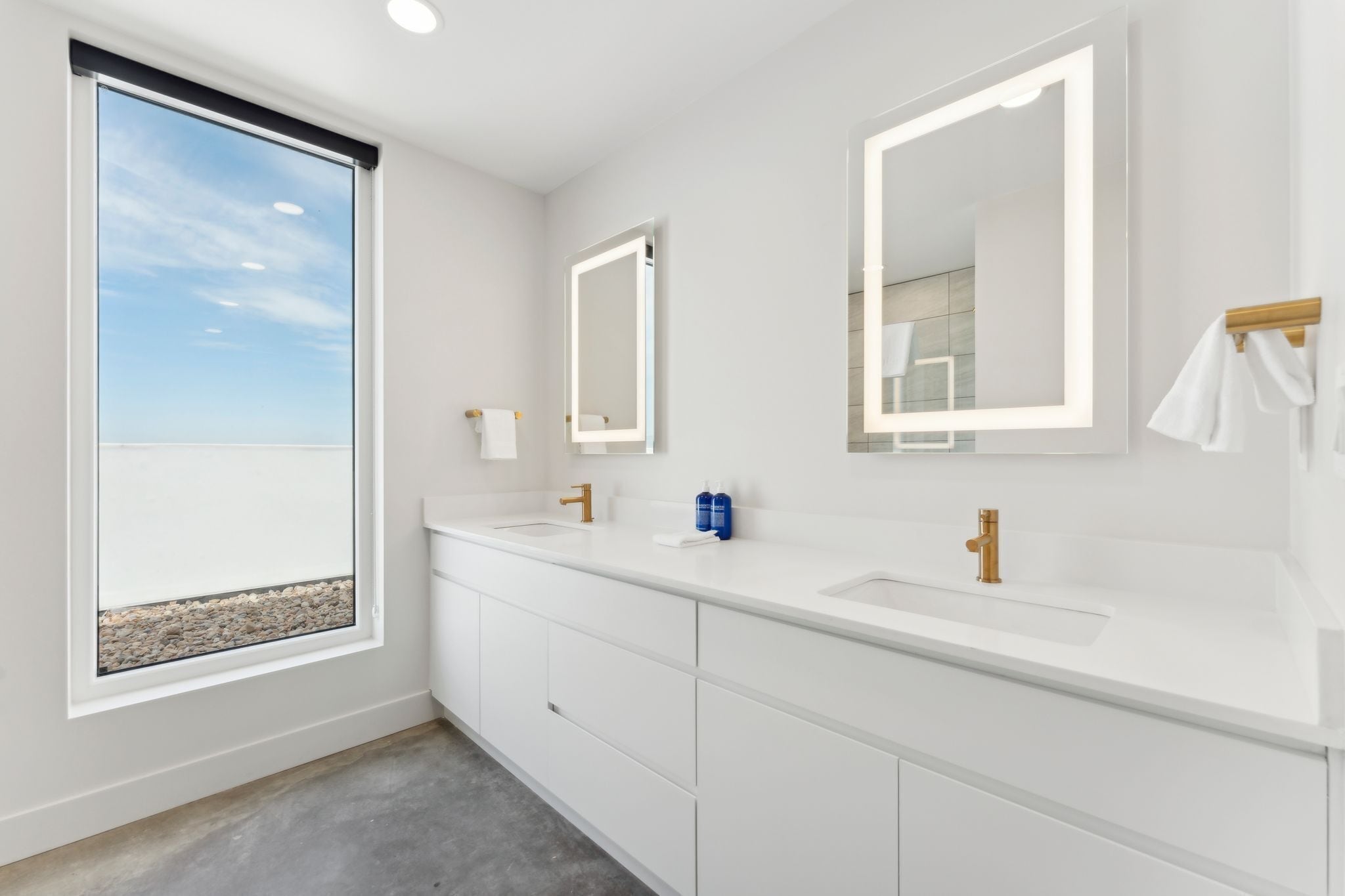A modern bathroom with two sinks, large mirrors, and a window with a view of the beach.