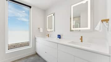 A modern bathroom with two sinks, large mirrors, and a window with a view of the beach.
