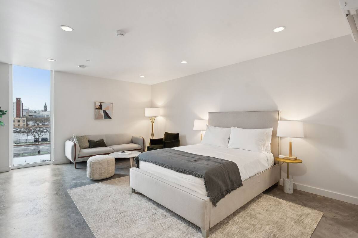 A modern bedroom with a large bed, bedside tables with lamps, a sofa, and a view of the city.