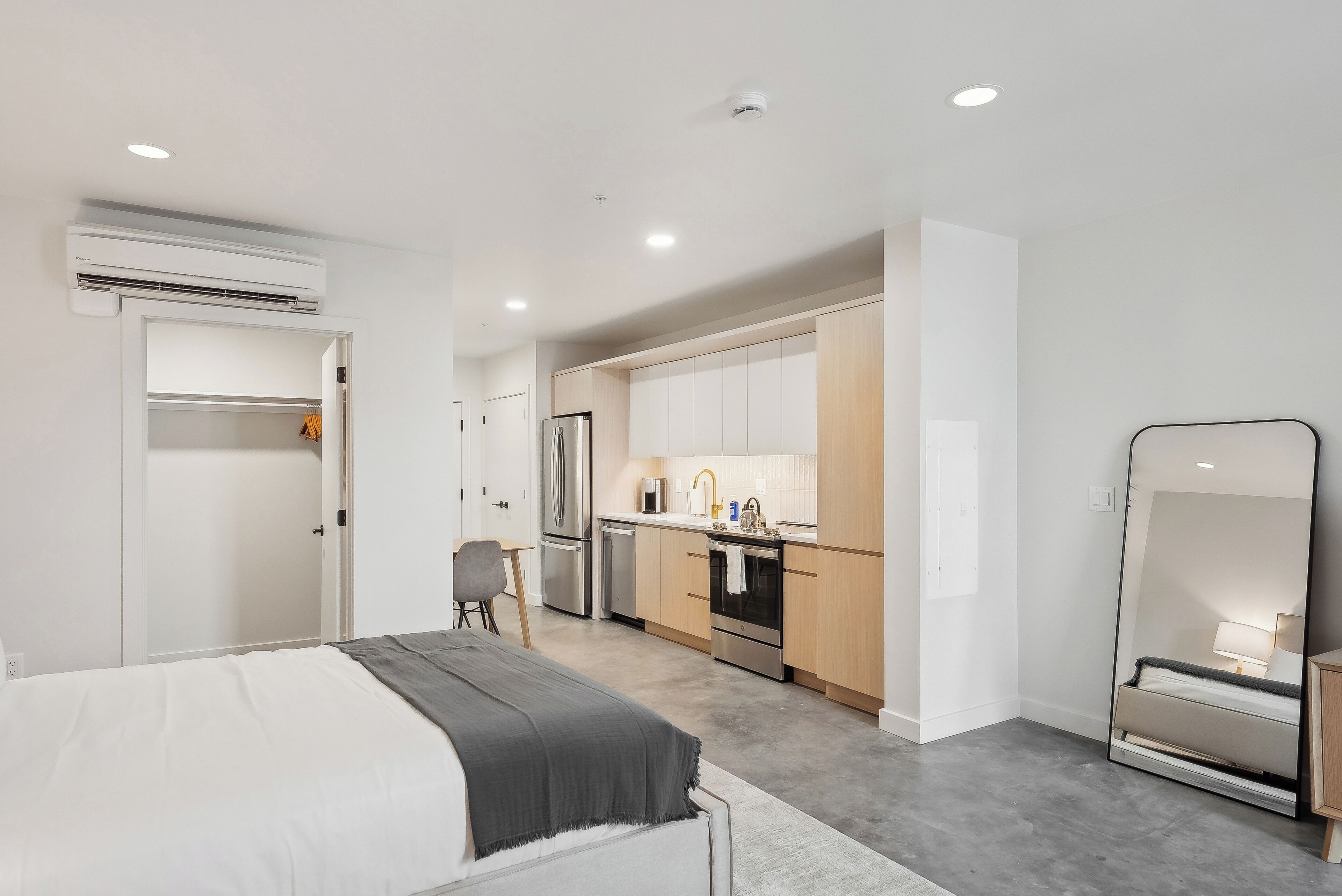 A modern bedroom with a bed, a small desk, and a chair. Adjacent is a compact kitchenette with a refrigerator, oven, and sink.