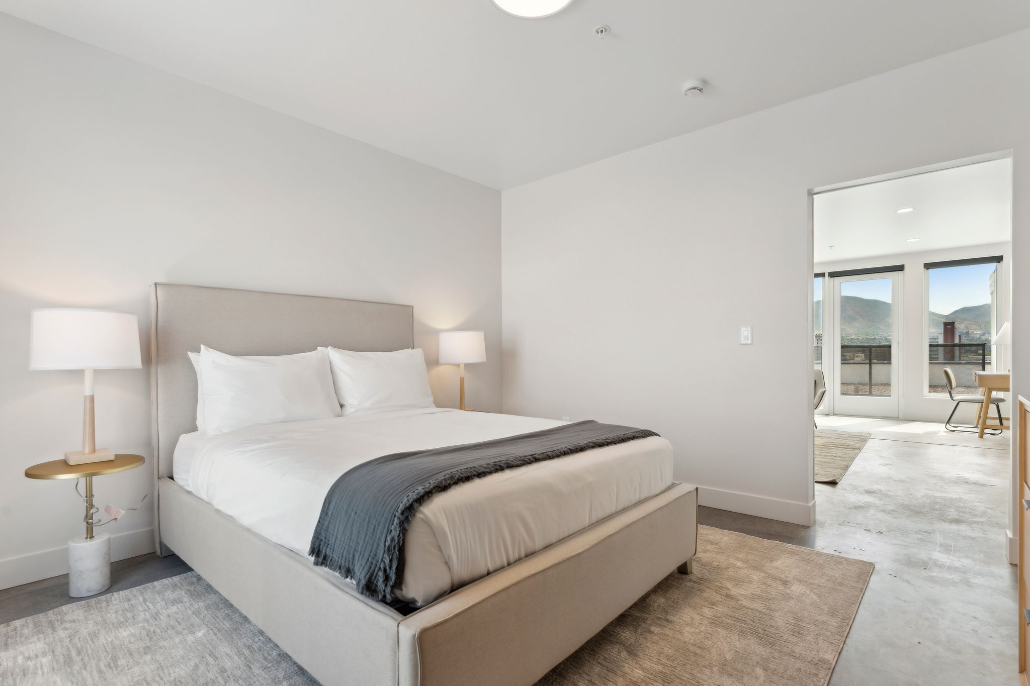 A modern hotel room with a large bed, two bedside lamps, a view of the outdoors through a sliding door, and a small table with a lamp.
