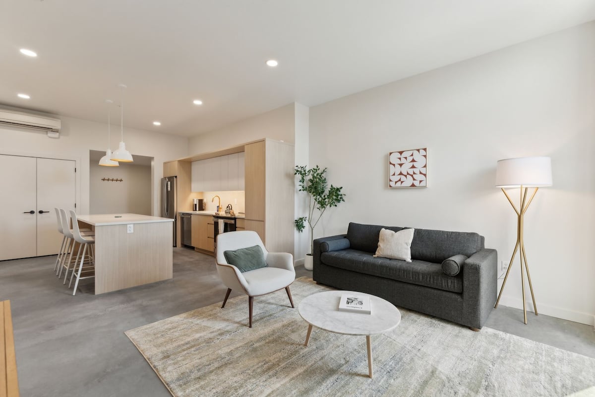 A modern living room with a grey sofa, a white armchair, and a round coffee table. There is a kitchen in the background with a dining area.