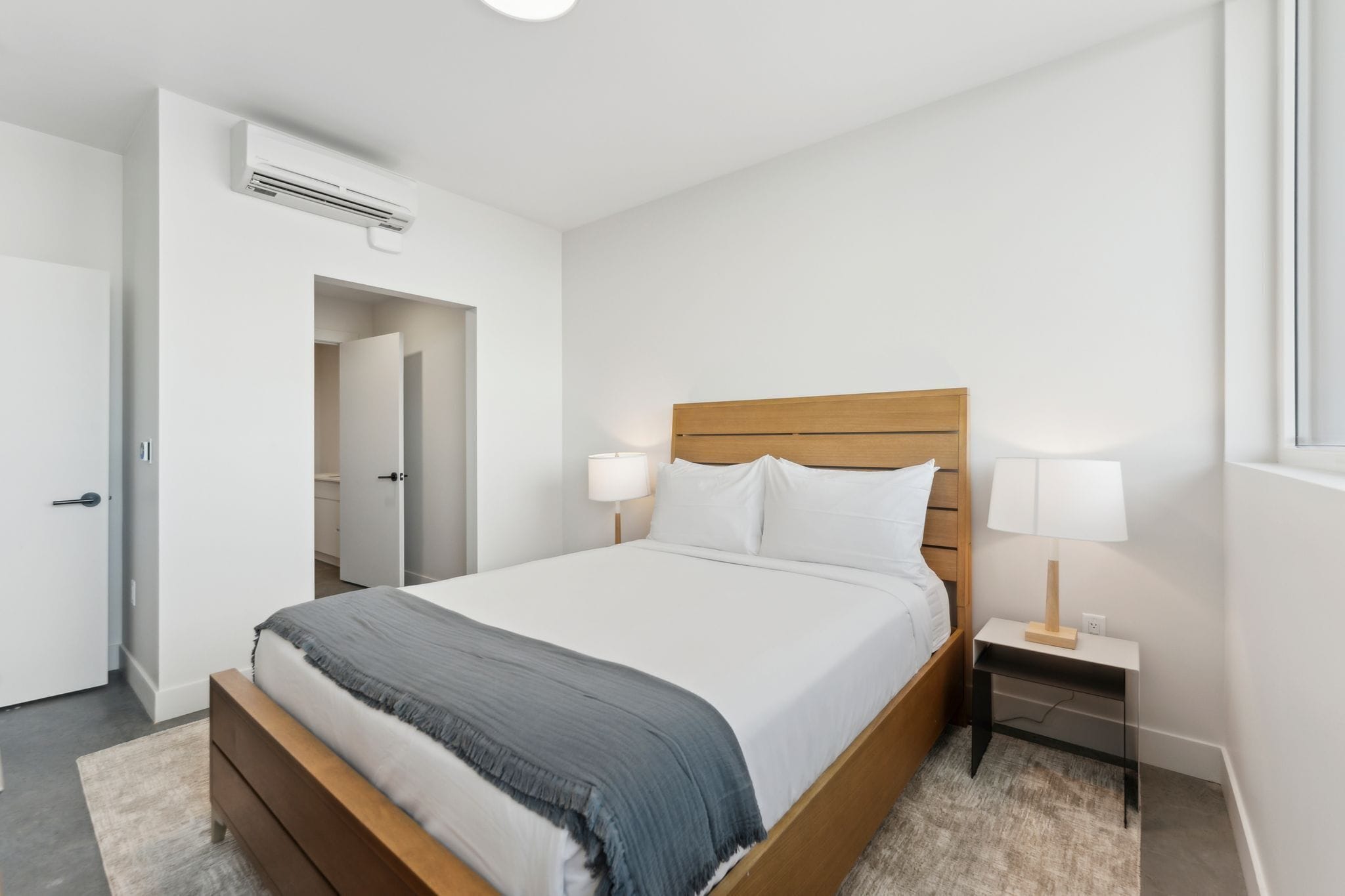 Un dormitorio con cama de madera, ropa de cama blanca, una mesita de noche con lámpara y un equipo de aire acondicionado.