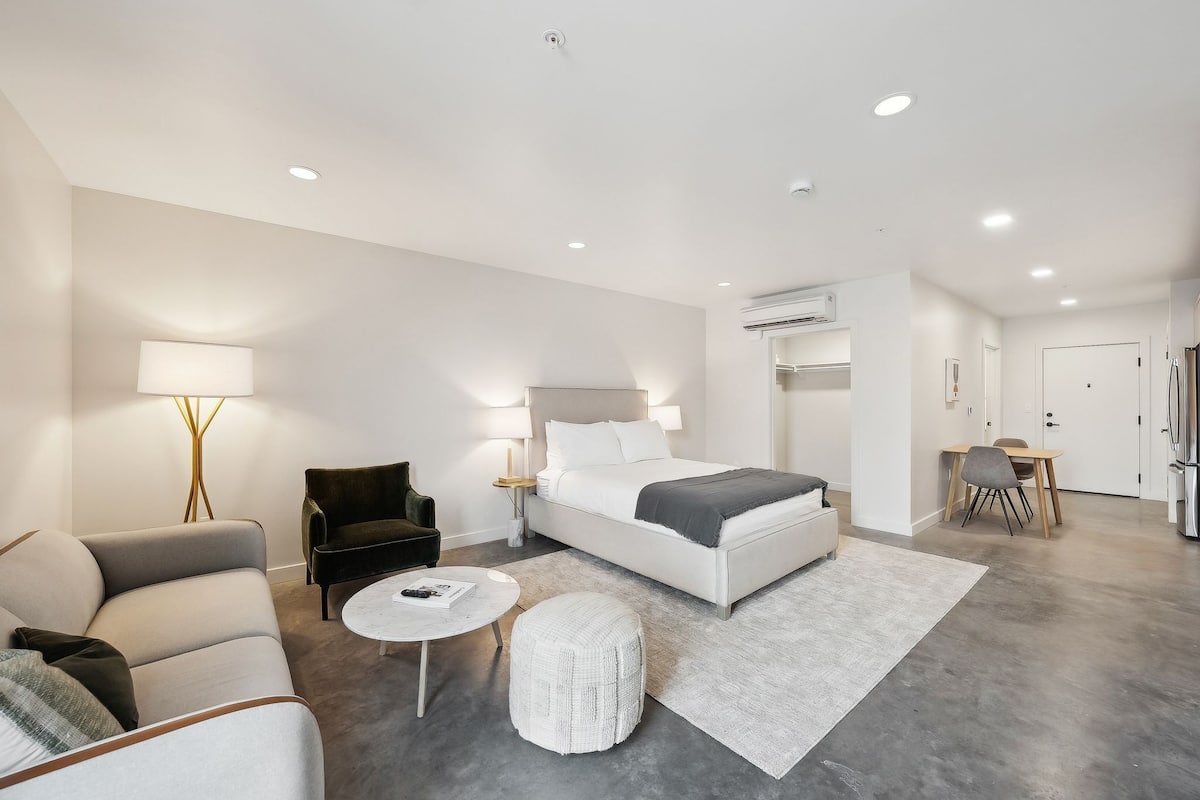 A modern hotel room with a large bed, a sofa, a small round table, and a dining area with chairs.