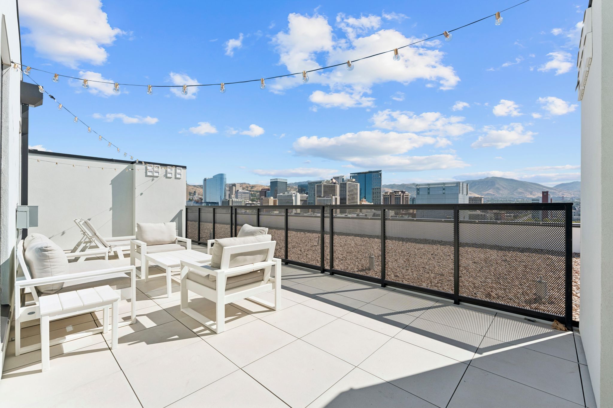 A rooftop terrace with white outdoor furniture, a string of lights, and a cityscape view.