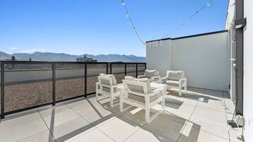 A rooftop terrace with white furniture, a view of mountains, and string lights.