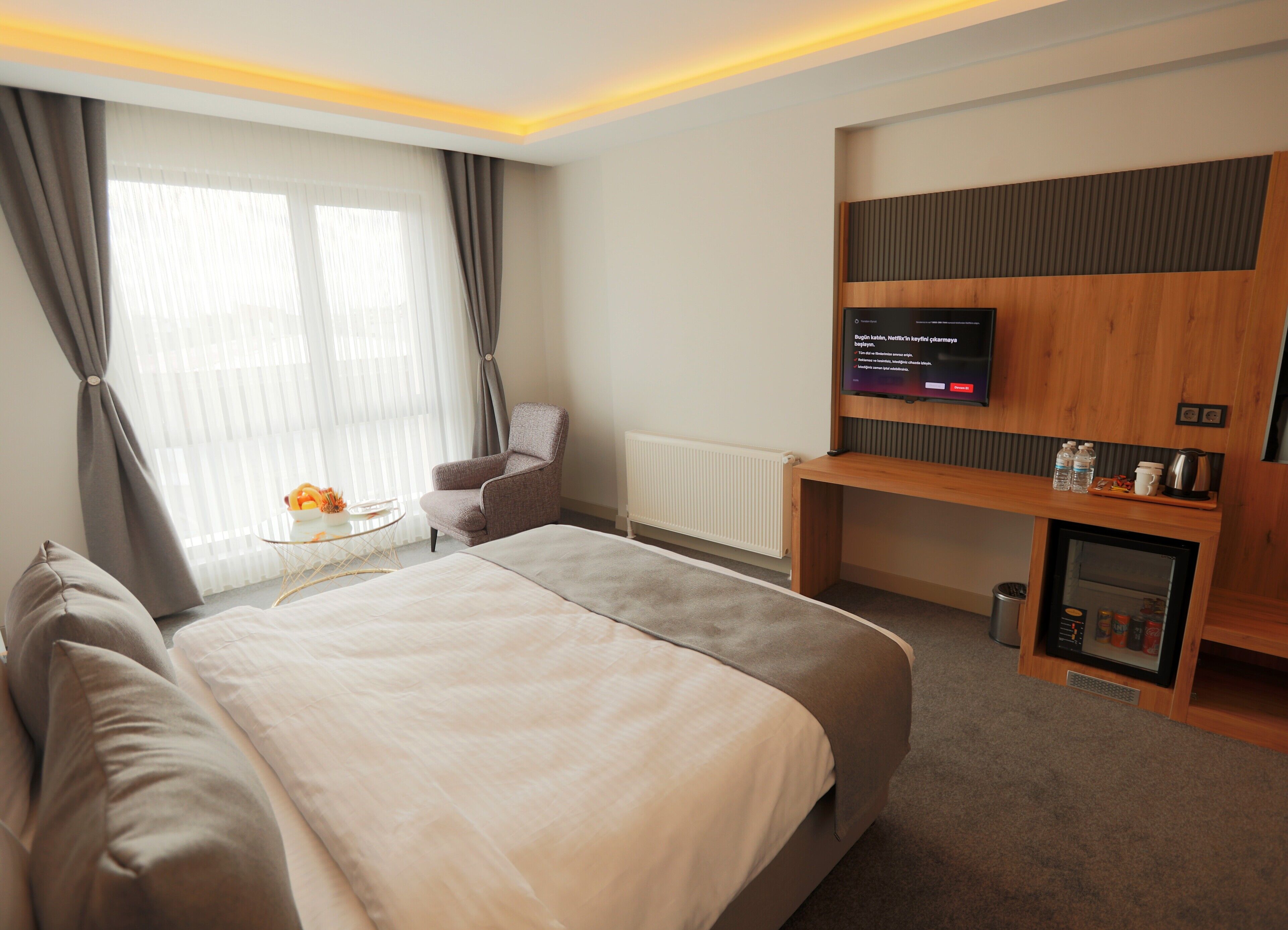A hotel room with a large bed, a chair, a TV, a small table with a fruit bowl, and a minibar.